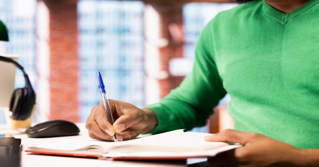 A man in a green sweater writes in a notebook, focusing on script translation of multi-language videos.