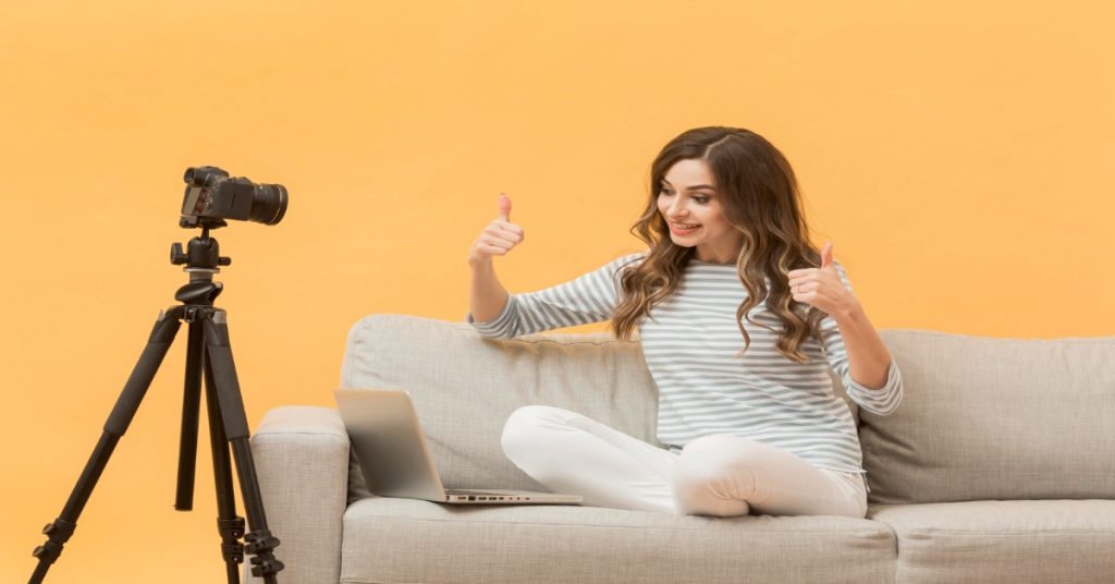 A woman on a couch using a laptop and camera for remote video production.